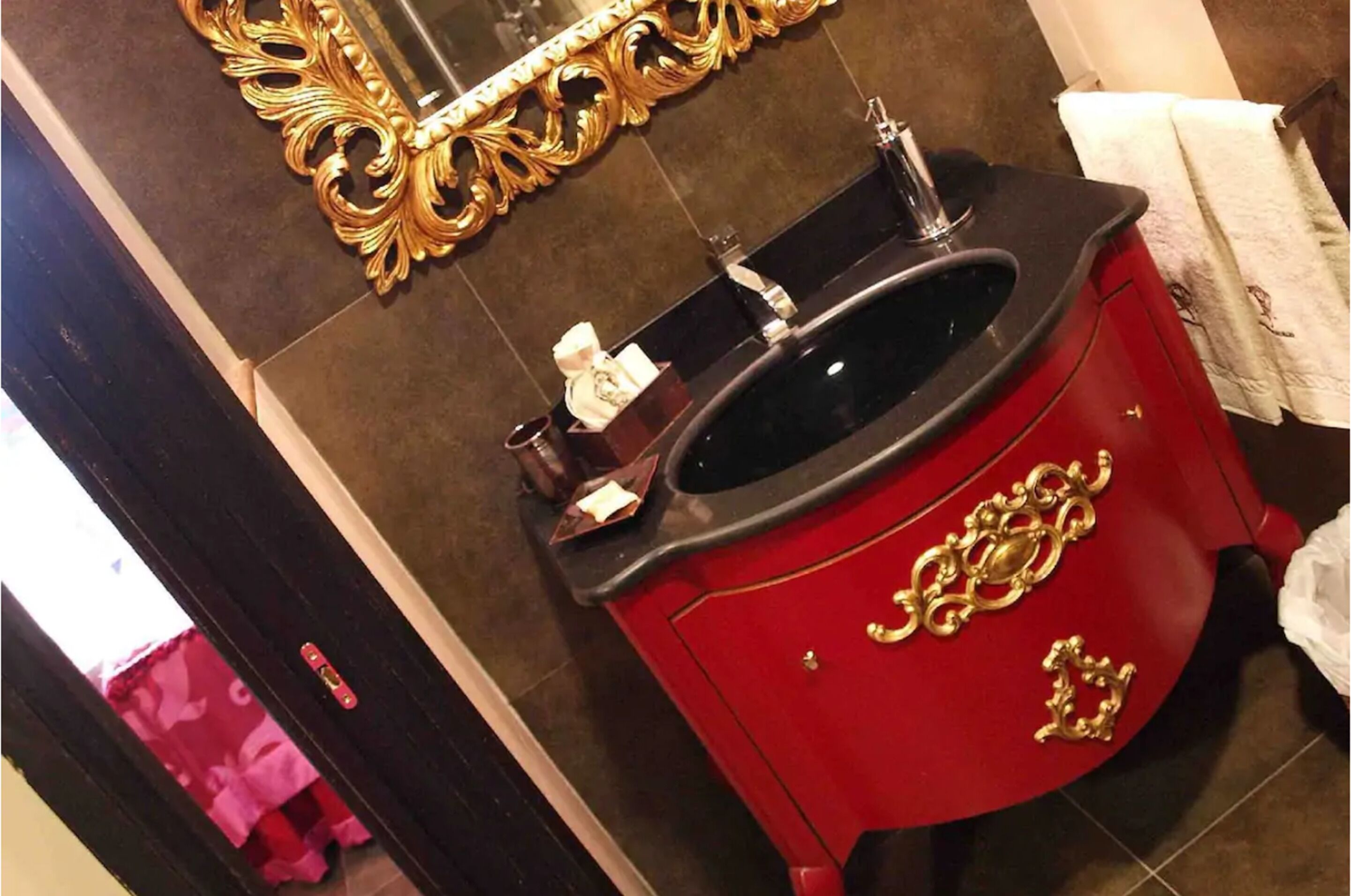 Un baño con tocador rojo, manijas doradas, lavabo negro y espejo con marco dorado.
