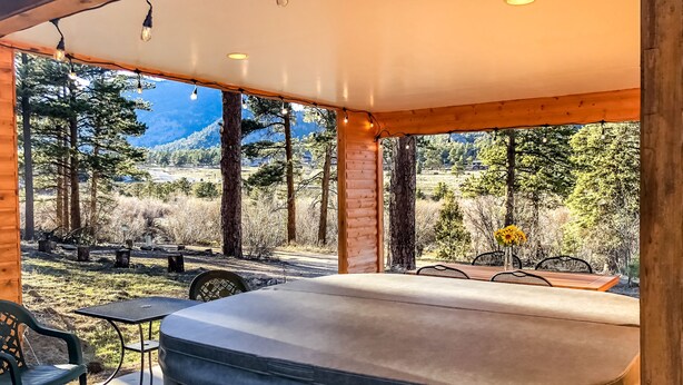 A covered outdoor seating area with a hot tub, wooden tables, and chairs, overlooking a natural landscape with trees and mountains. - Mountain Cabin Overlooking Breathtaking Riverfront and Valley Views  (Estes Park)