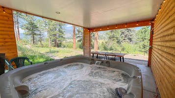 Un bain à remous avec une table et des chaises, situé à l’extérieur d’un chalet en bois, entouré d’arbres et offrant une vue dégagée sur le paysage.
