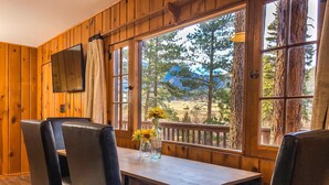 A wooden cabin interior with a dining table, chairs, and a window offering a view of trees and mountains. - Mountain Cabin Overlooking Breathtaking Riverfront and Valley Views  (Estes Park)