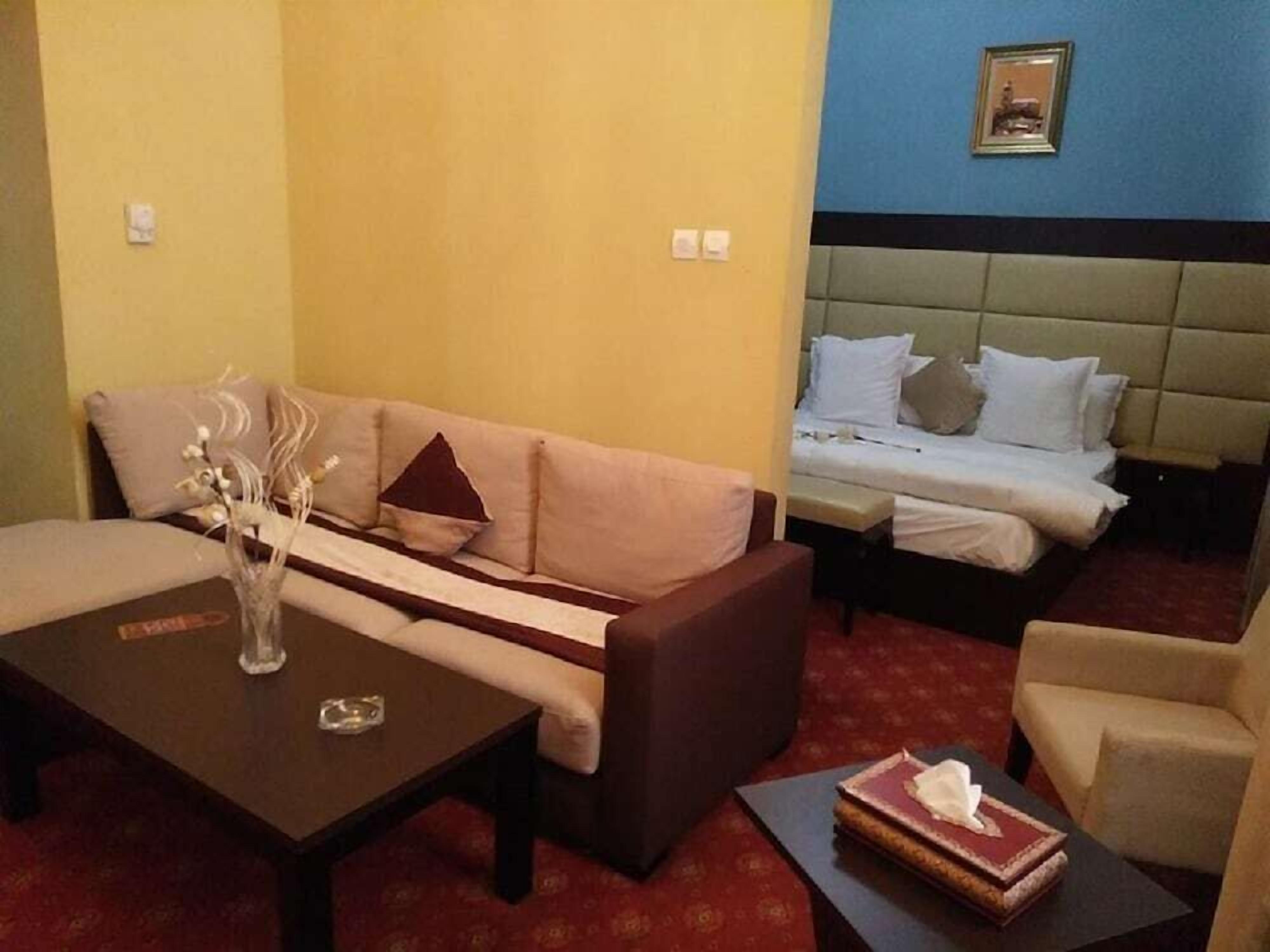 A hotel room with a sofa, a small table with a vase, a bed with pillows, and a chair.