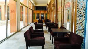 Un restaurante con mesas y sillas dispuestas en fila, grandes ventanales en un lado y una pared con diseño decorativo en el otro.