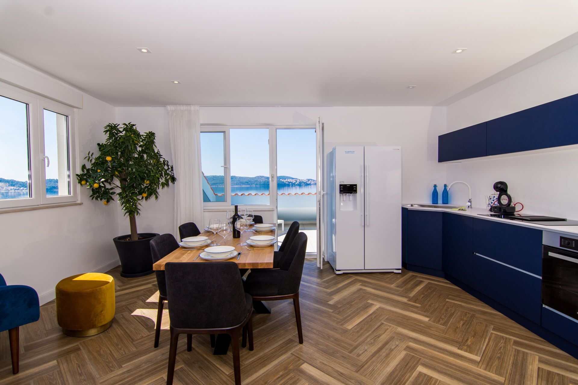 Une cuisine moderne dotée d’un îlot central intégré, un coin repas avec une table et des chaises en bois, et une vue sur la mer grâce à de grandes fenêtres.