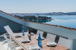 A dining table set with plates, glasses, and bottles, overlooking a coastal view with a clear sky. - Apartment Deep Blue near Trogir (Trogir)