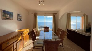A dining area with a glass table and wicker chairs, a wooden cabinet, and a view of the sea through large windows. - Ático Vistamar X2113 - Benitachell, Costa Blanca (Benitachell)