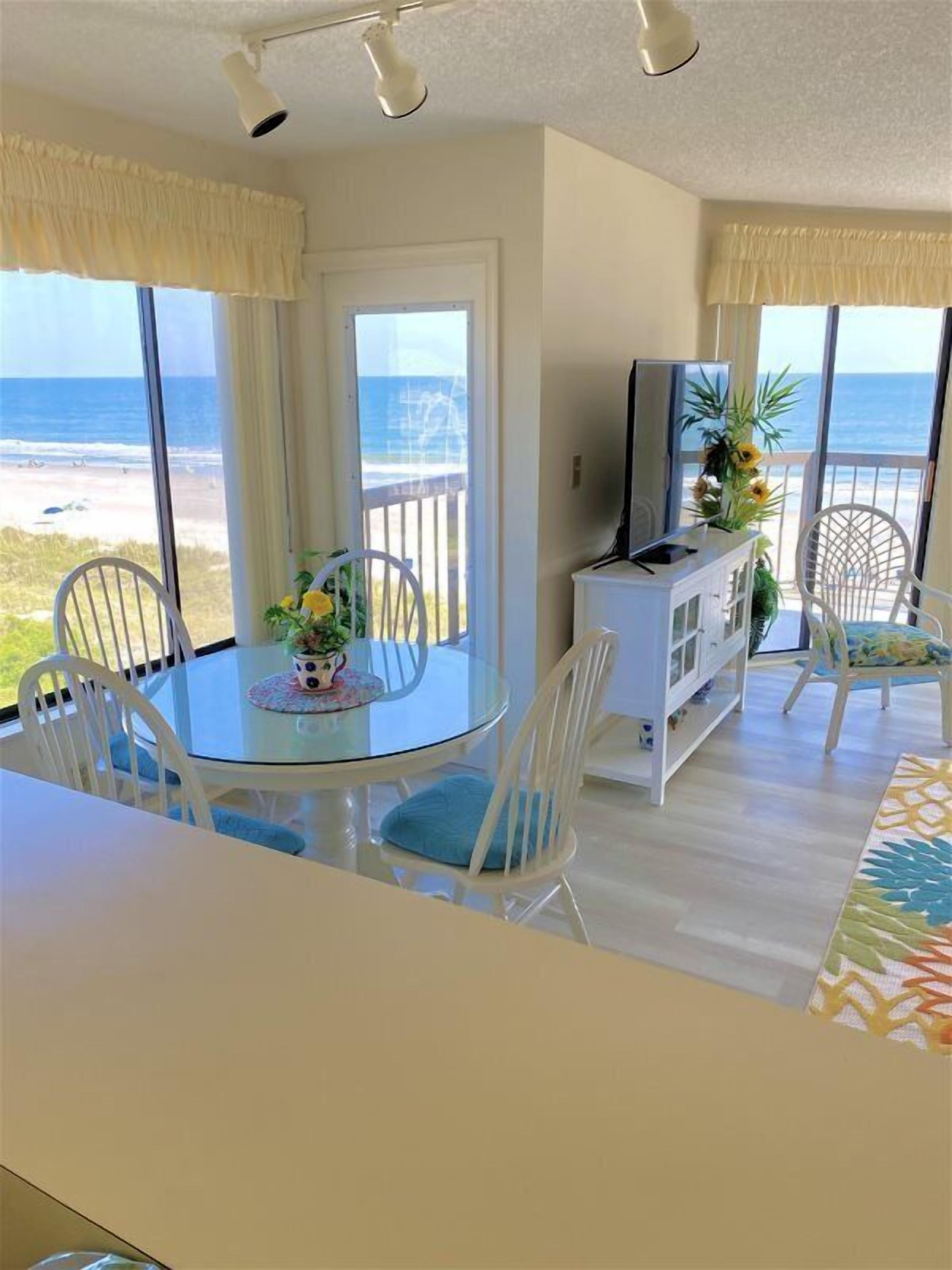 A room with a glass table, chairs, a TV, and a view of the beach.