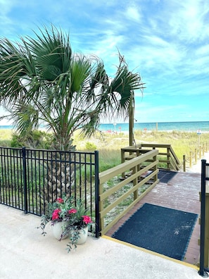A wooden pathway leading to a beach with a metal gate, a palm tree, and a potted plant with pink flowers.