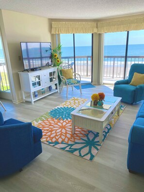 A living room with a TV, a white cabinet, a blue sofa, a blue armchair, a patterned rug, and a balcony with a view of the ocean.