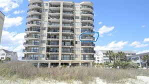 Un hôtel à plusieurs étages, doté de balcons à chaque niveau, situé près d’une plage bordée de dunes de sable.
