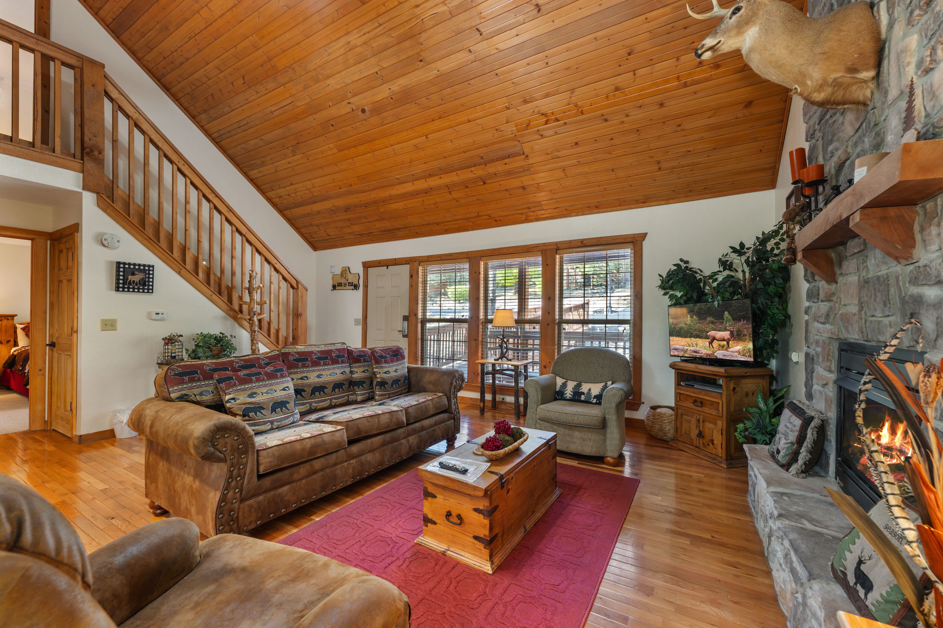 A spacious living room with a wooden ceiling, a stone fireplace, a large television, and a staircase leading to an upper level.