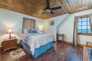 Une chambre à coucher avec un lit, une table de chevet, une lampe, un ventilateur de plafond, une fenêtre avec des rideaux et une vue sur la mer.