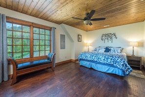 A bedroom with a wooden ceiling, a large window with a bench, a bed with a blue floral bedspread, and two bedside lamps.