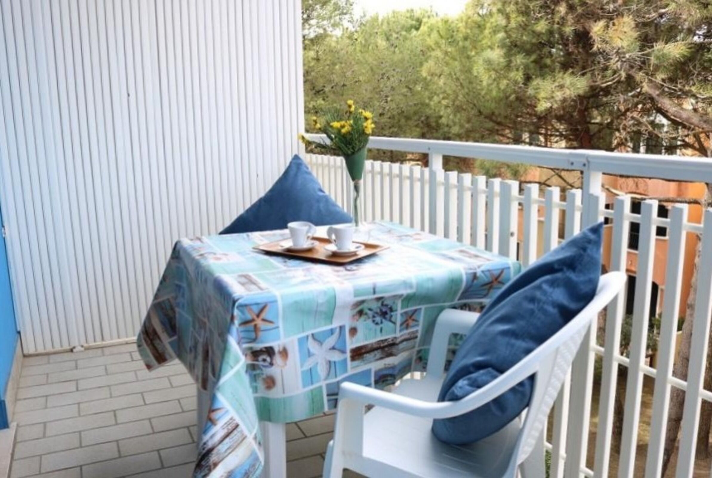 Una pequeña mesa exterior con mantel azul, sillas blancas y valla blanca.