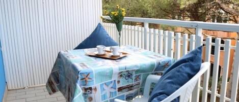 Una pequeña mesa exterior con mantel azul, sillas blancas y valla blanca.