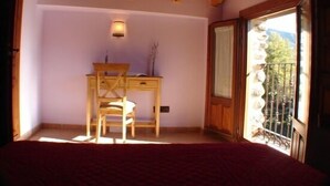 Un dormitorio con un escritorio y silla de madera, una cama y un balcón con vistas a árboles y montañas.