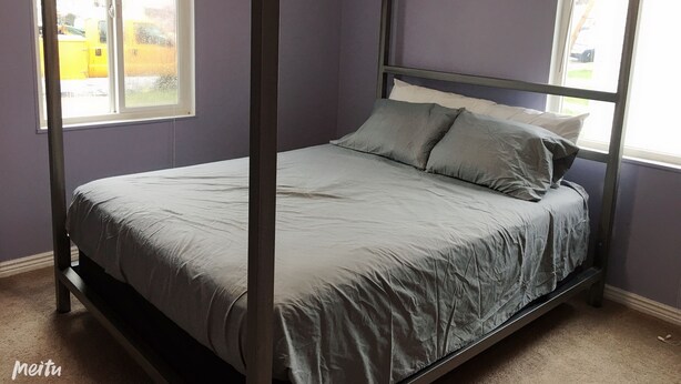 Uma cama com dossel, roupa de cama cinzenta, num quarto com paredes roxas e duas janelas.