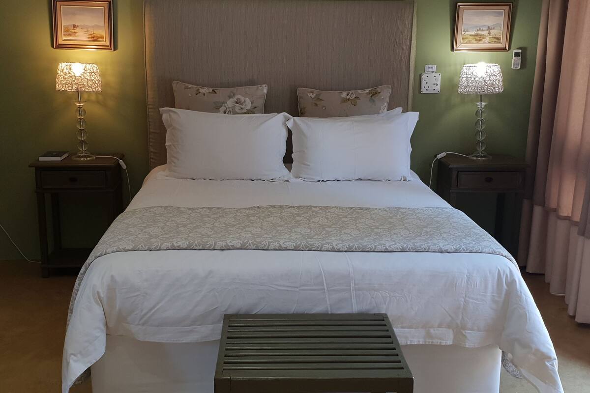 Una cama bien hecha con sábanas blancas y un plaid estampado, flanqueada por dos mesitas de noche con lámparas, en una habitación con paredes verdes y una ventana con cortinas.