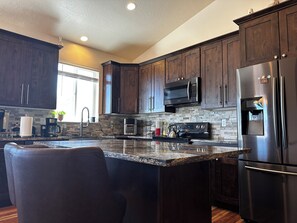 A modern kitchen with dark wood cabinets, a stainless steel refrigerator, a built-in microwave, and a granite countertop. - Hot Tub, Spacious 6BR Home w 2 Large Living Areas & 3 Bath (Murray)