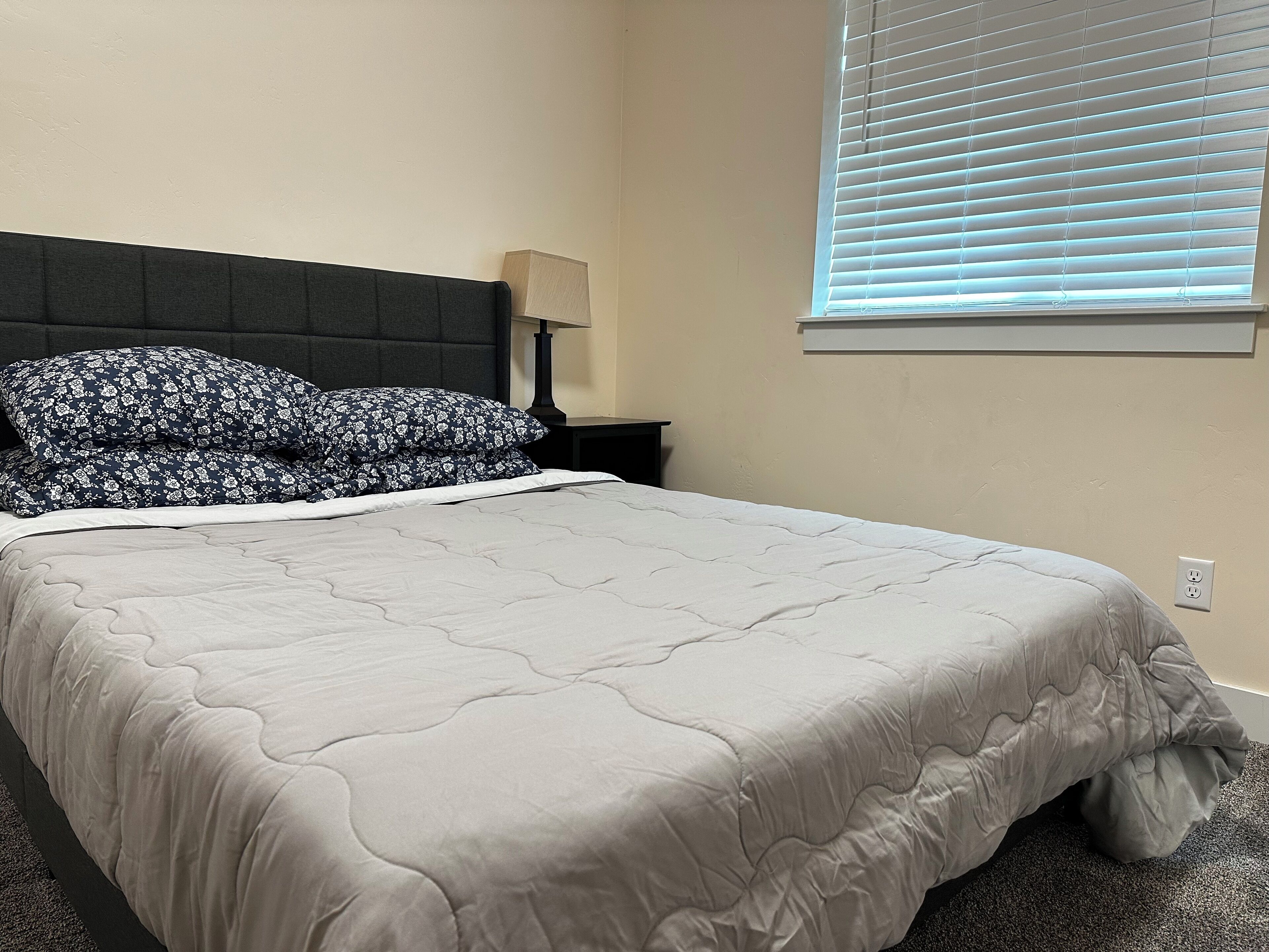 A neatly made bed with a grey quilt and floral pillows, a bedside table with a lamp, and a window with blinds.