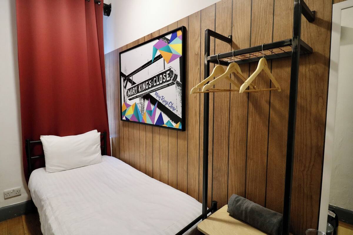 A bedroom with a bed, a wooden wall panel, a colorful artwork, and a clothing rack.