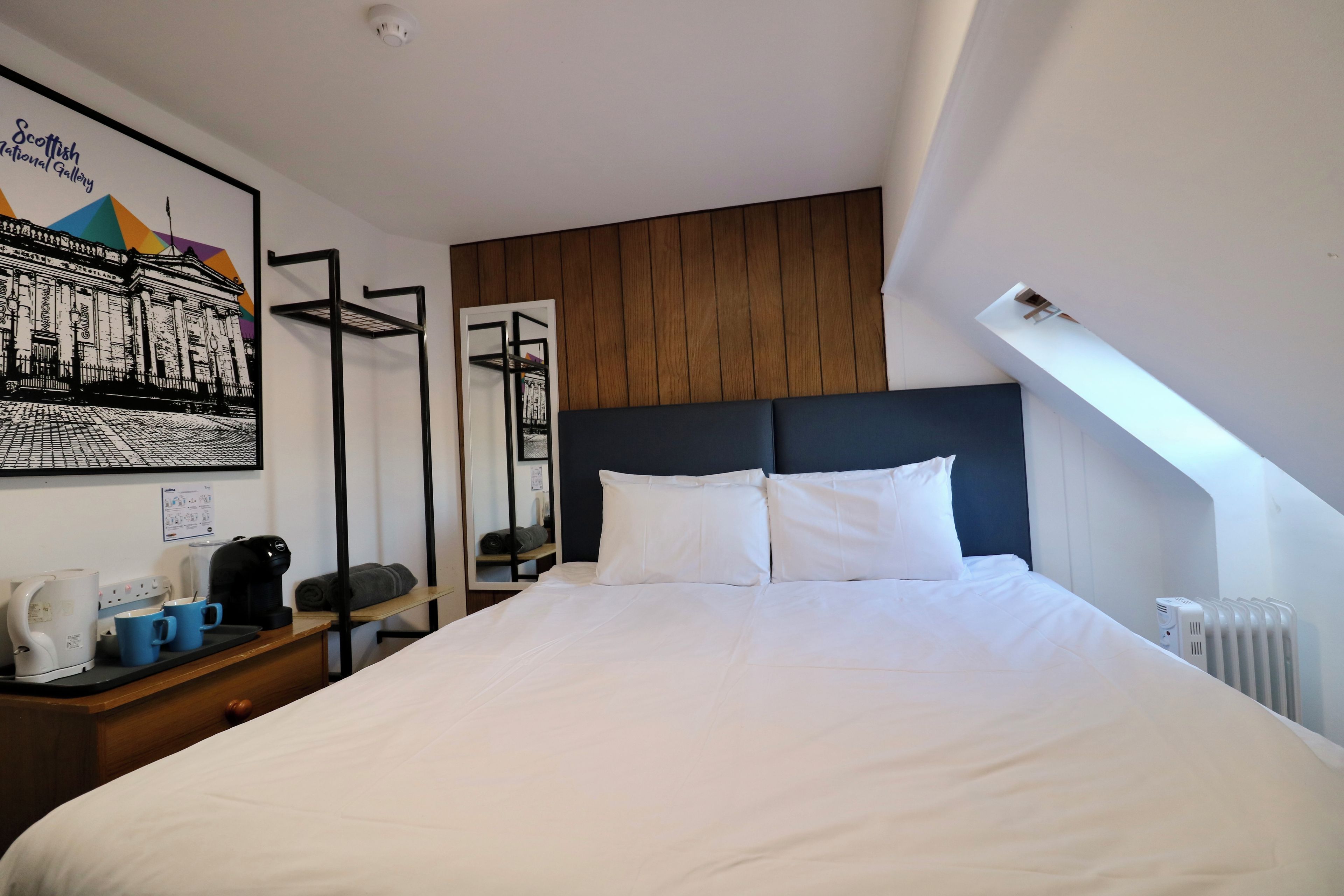 Une chambre d’hôtel avec un grand lit, une tête de lit en bois, un mur blanc orné d’un tableau encadré, une petite table avec une bouilloire et des tasses, et un radiateur.