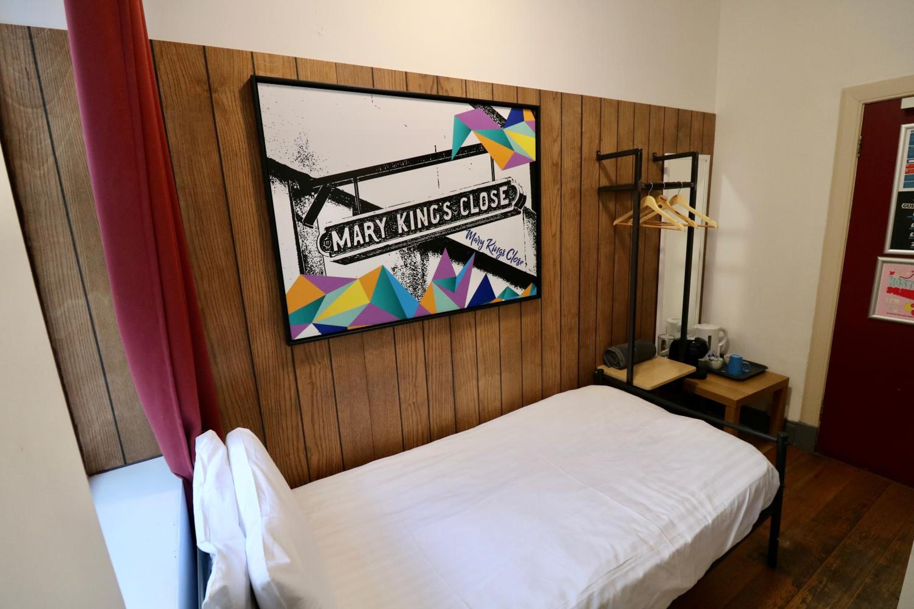 A single bed with a white sheet, a wooden headboard, a wall-mounted artwork with geometric shapes and text, a small wooden side table with a cup and a book, and a red curtain partially visible.
