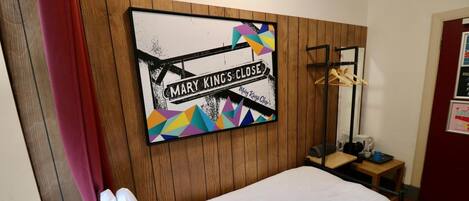 A single bed with a white sheet, a wooden headboard, a wall-mounted artwork with geometric shapes and text, a small wooden side table with a cup and a book, and a red curtain partially visible.