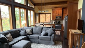 A modern kitchen with wooden cabinets, a dining area with a gray sectional sofa, and a large window offering a view of trees.