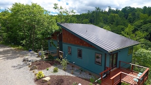 A small, single-story cabin with a metal roof, surrounded by a gravel driveway and a landscaped garden.