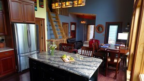 A kitchen with granite countertops, a stainless steel refrigerator, wooden cabinets, a dining area with a table and chairs, a staircase, and a living room with a sofa and a TV.