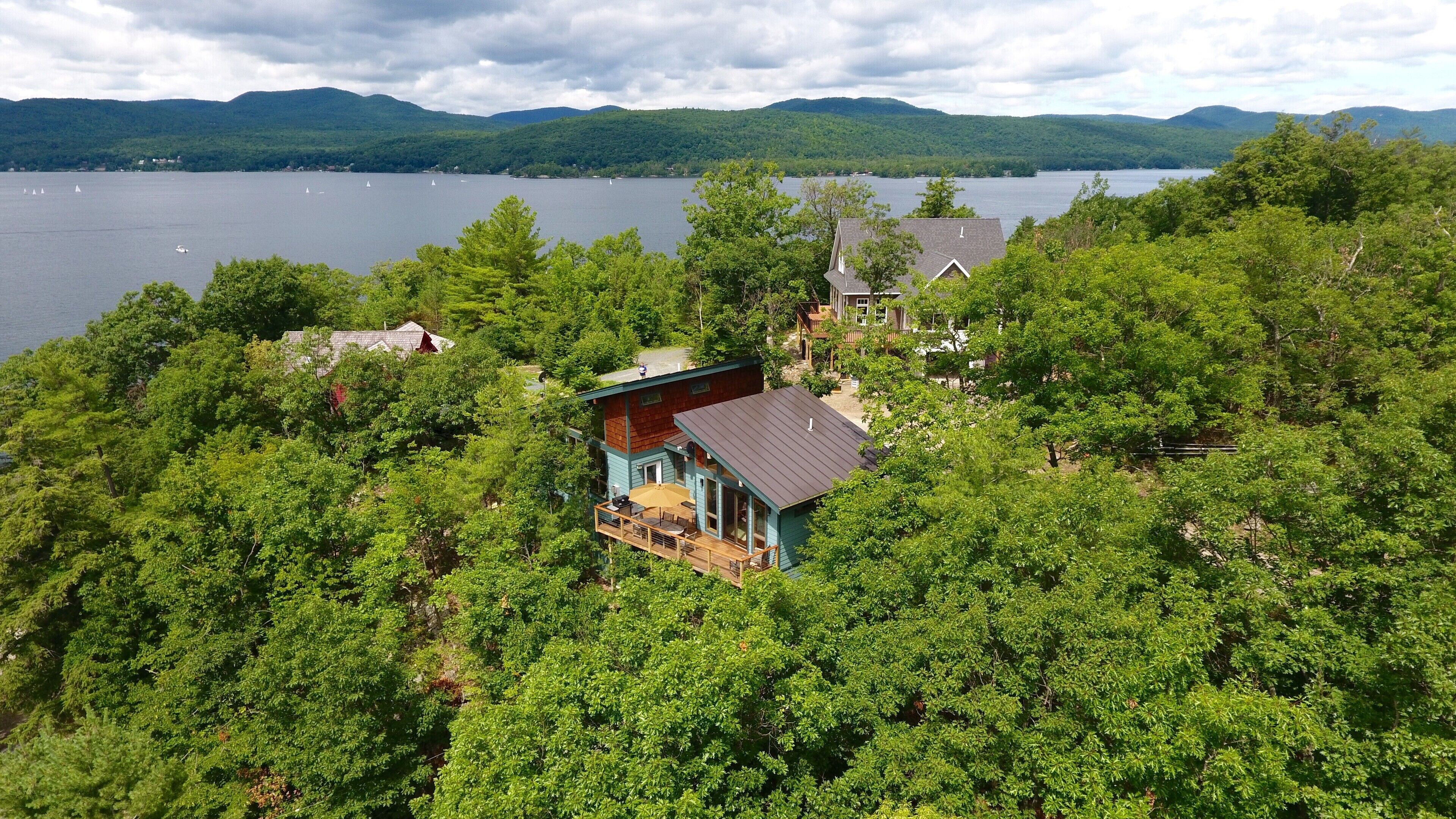 A lakeside cabin with a deck, surrounded by dense forest, overlooking a lake with sailboats.