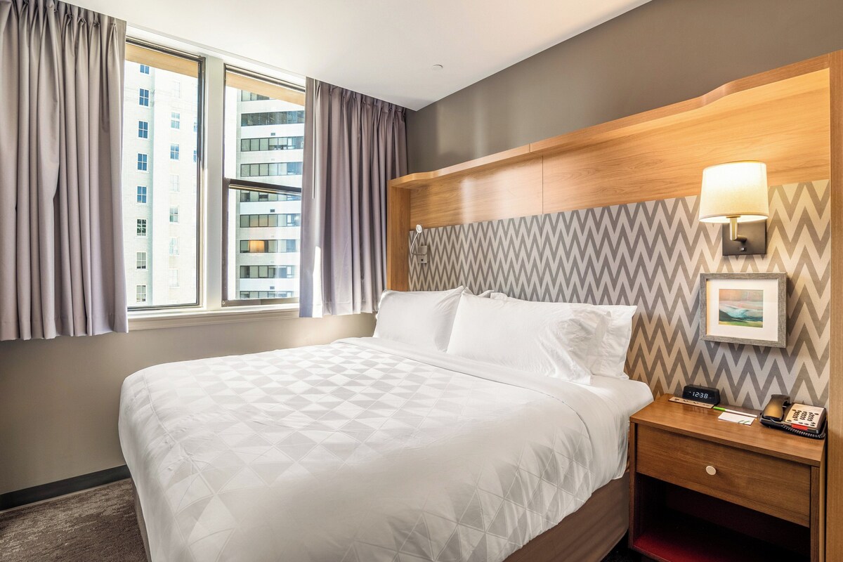 A hotel room with a large bed, a wooden headboard, a nightstand with a lamp, and a view of buildings through the window.