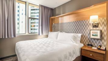 Ein Hotelzimmer mit einem großen Bett, einem hölzernen Kopfteil, einem Nachttisch mit Lampe und Blick auf Gebäude durch das Fenster.