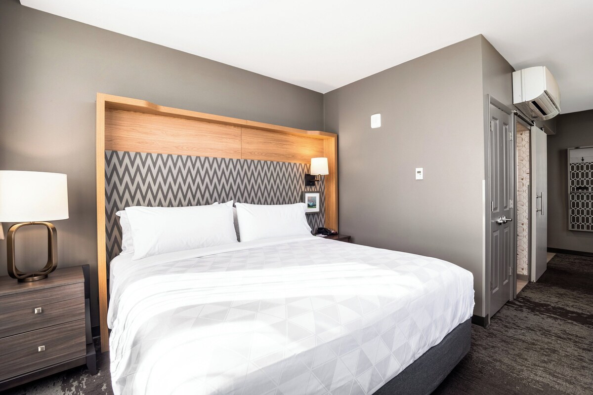A hotel room with a large bed, a wooden headboard, a nightstand with a lamp, and a door leading to another room.