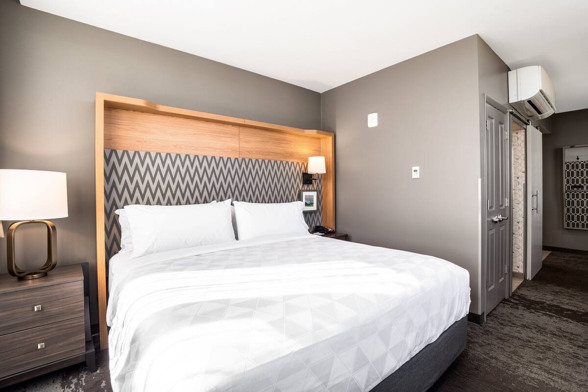 A hotel room with a large bed, a wooden headboard, a nightstand with a lamp, and a door leading to another room.