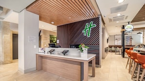 A reception area with a wooden reception desk, a sign with a green logo, a digital phone, and a plant. - Holiday Inn Columbia - Downtown by IHG (Columbia)