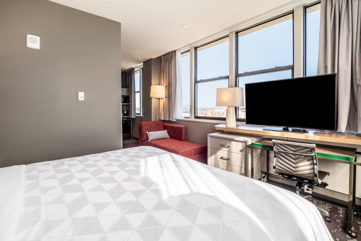 A hotel room with a large bed, a desk with a flat-screen TV, a chair, and a window with curtains.
