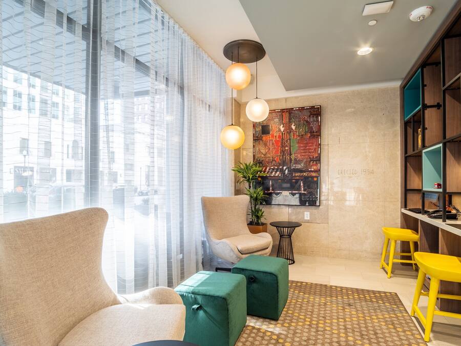 A modern living room with a large window, a beige armchair, a green ottoman, yellow bar stools, and a painting on the wall.