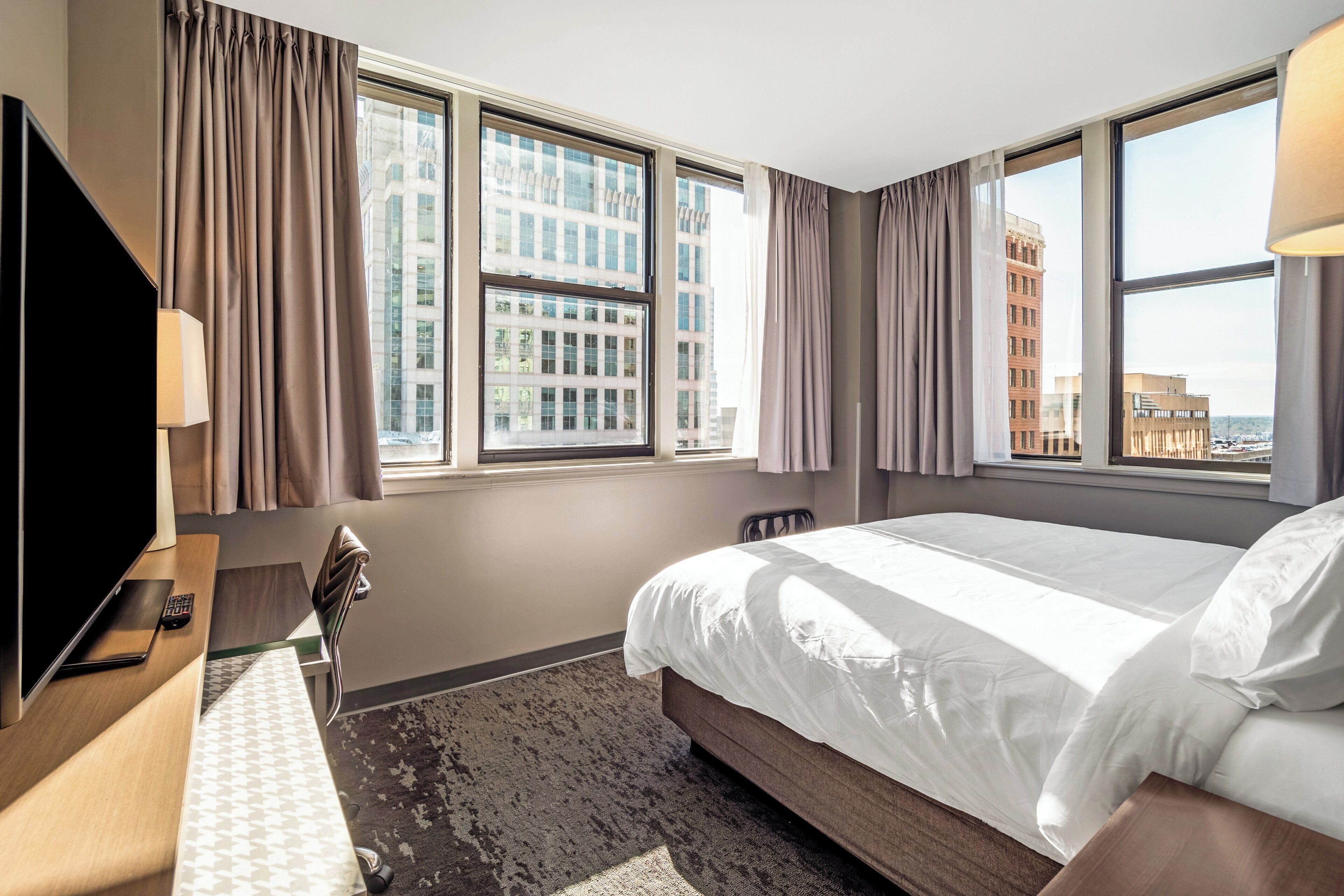 Een hotelkamer met een bed, een bureau met televisie, een stoel en grote ramen met gordijnen.