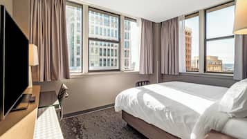 Een hotelkamer met een bed, een bureau met televisie, een stoel en grote ramen met gordijnen.