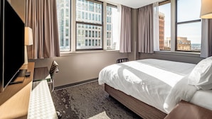 A hotel room with a large bed, a desk with a TV, and a view of buildings through the windows. - Holiday Inn Columbia - Downtown by IHG (Columbia)