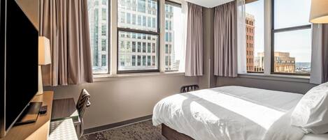 Quarto de hotel com uma cama grande, uma escrivaninha com televisão e vista para prédios pelas janelas.