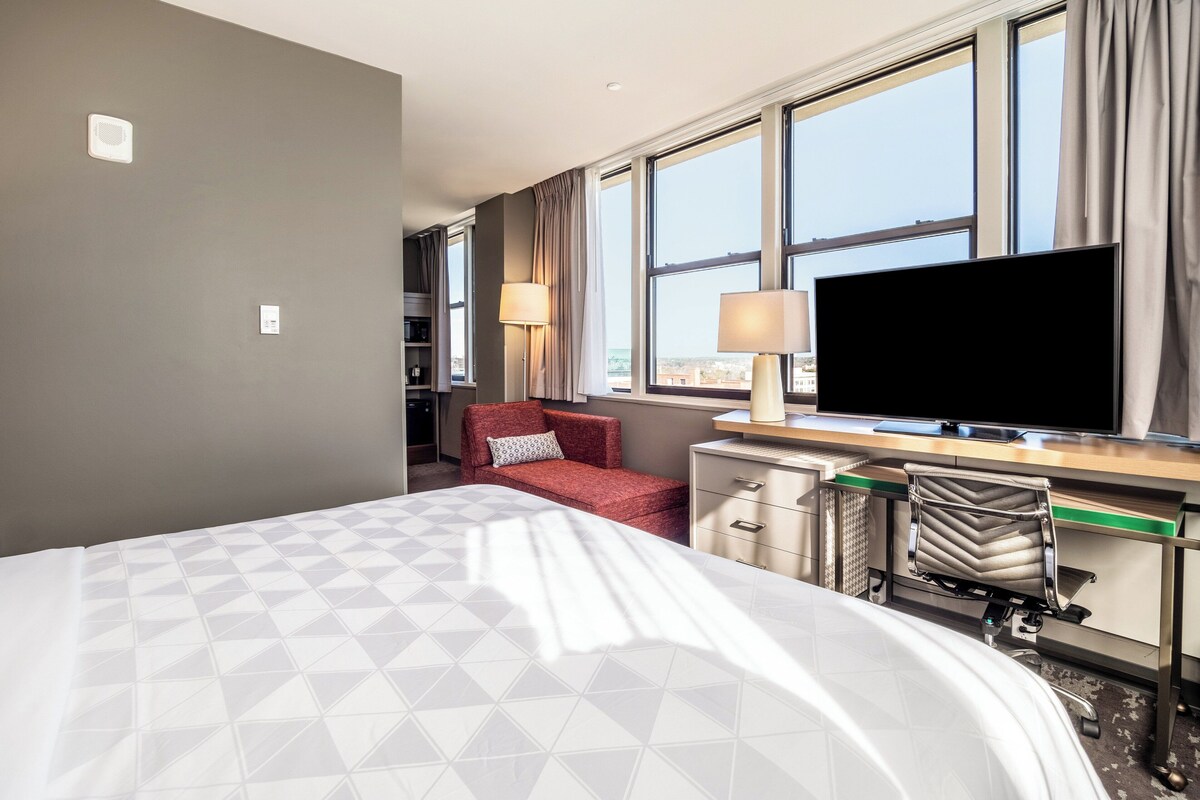 A hotel room with a large bed, a desk with a flat-screen TV, a chair, and a window with curtains.
