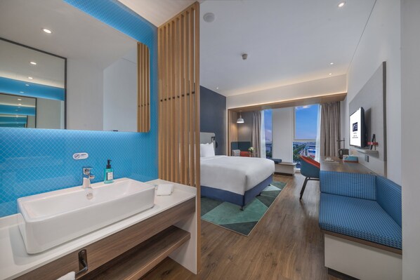A modern hotel room with a large white sink, a bed with blue bedding, a blue sofa, and a view of the cityscape. - Holiday Inn Express Qingdao Chengyang Central by IHG (Qingdao)