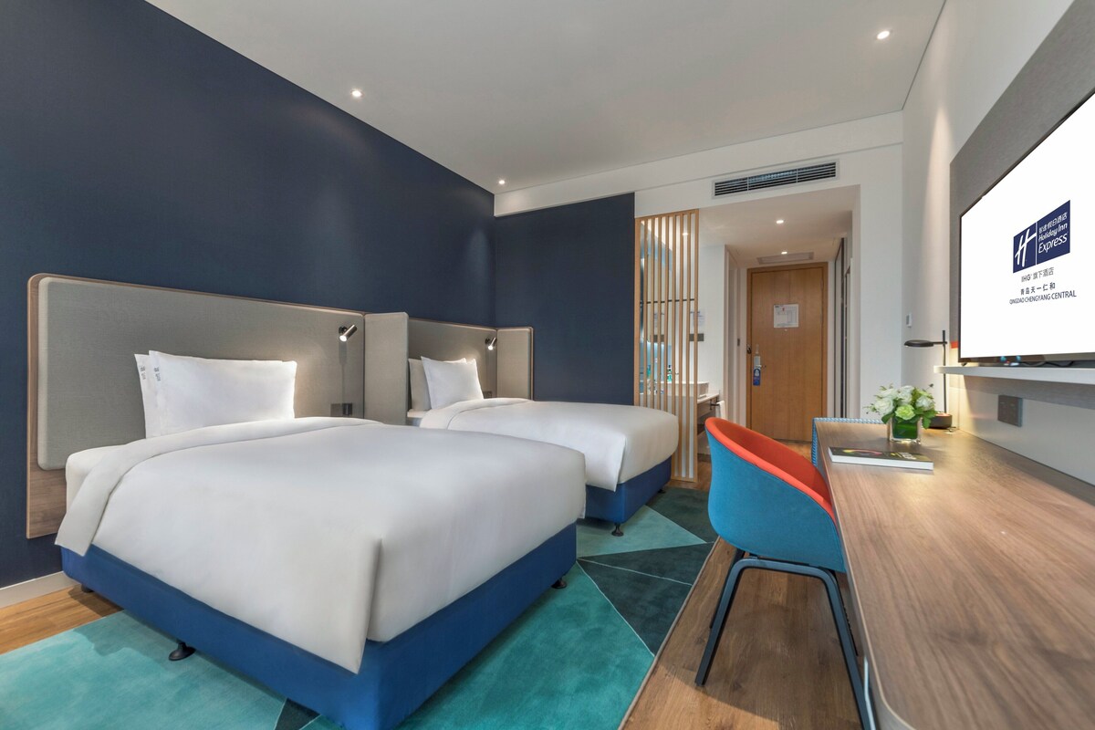 A hotel room with two beds, a desk, and a view of the hallway.