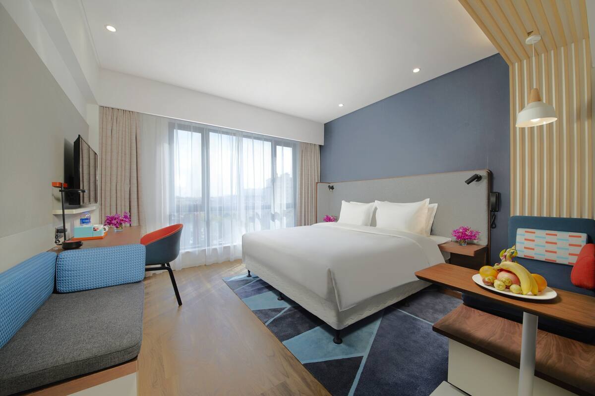 Ein modernes Hotelzimmer mit einem großen Bett, einem Schreibtisch mit Obstschale, einem blauen Sessel und einem Fernseher auf einem Standfuß.
