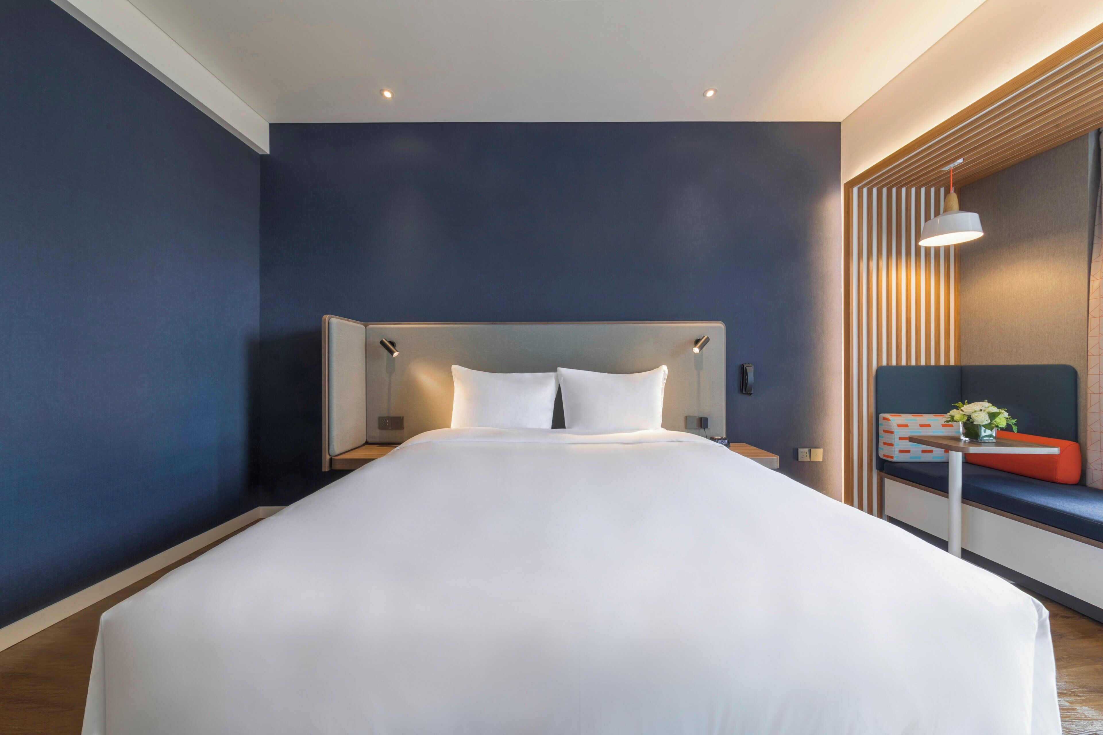 Una habitación de hotel moderna con una cama grande, una mesita de noche y una zona de estar con un banco y una pequeña mesa.