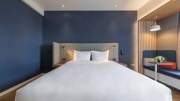 Una habitación de hotel moderna con una cama grande, una mesita de noche y una zona de estar con un banco y una pequeña mesa.