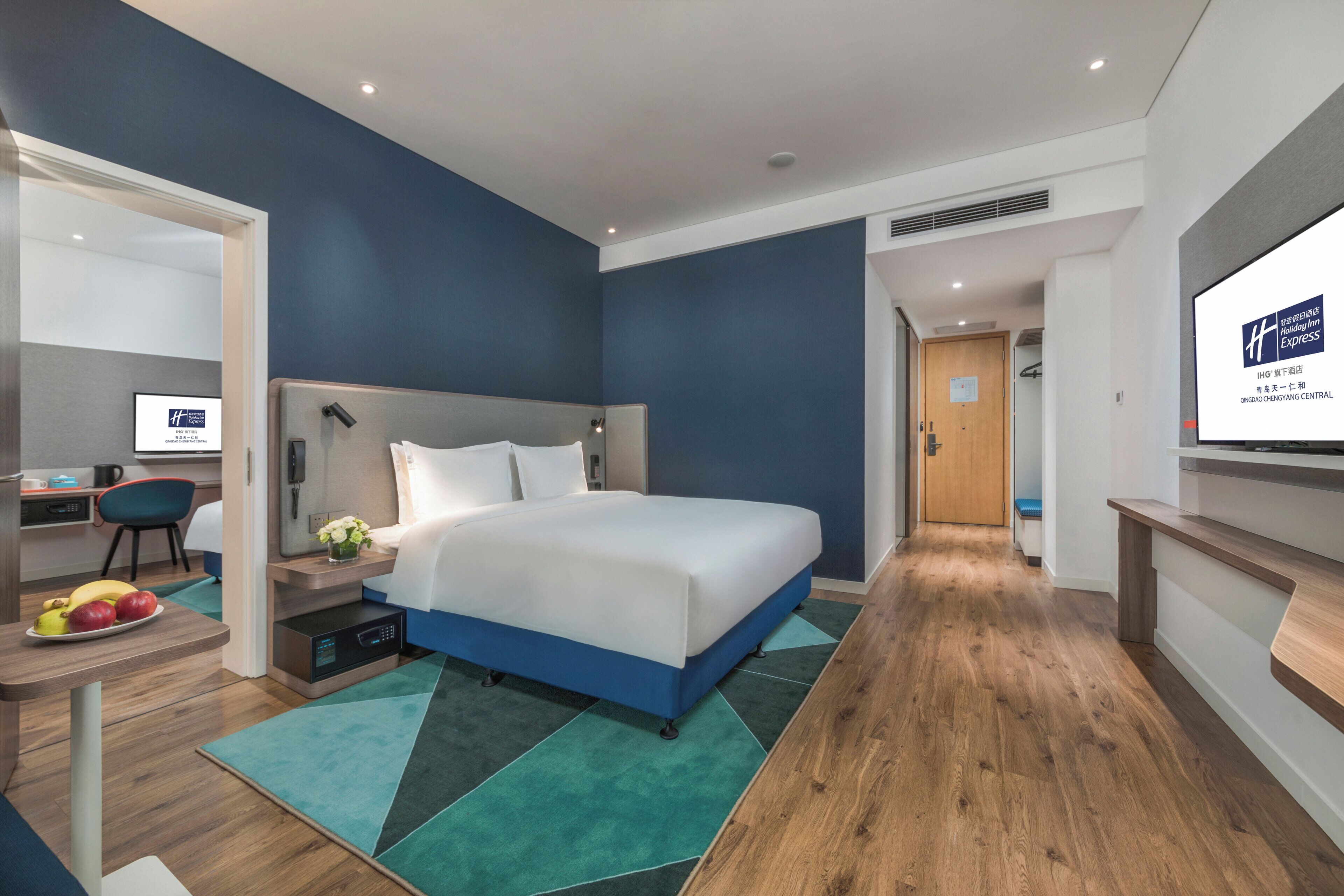 Habitación de hotel moderna con una cama grande, un escritorio con computadora y una pantalla de televisión mostrando información del hotel.