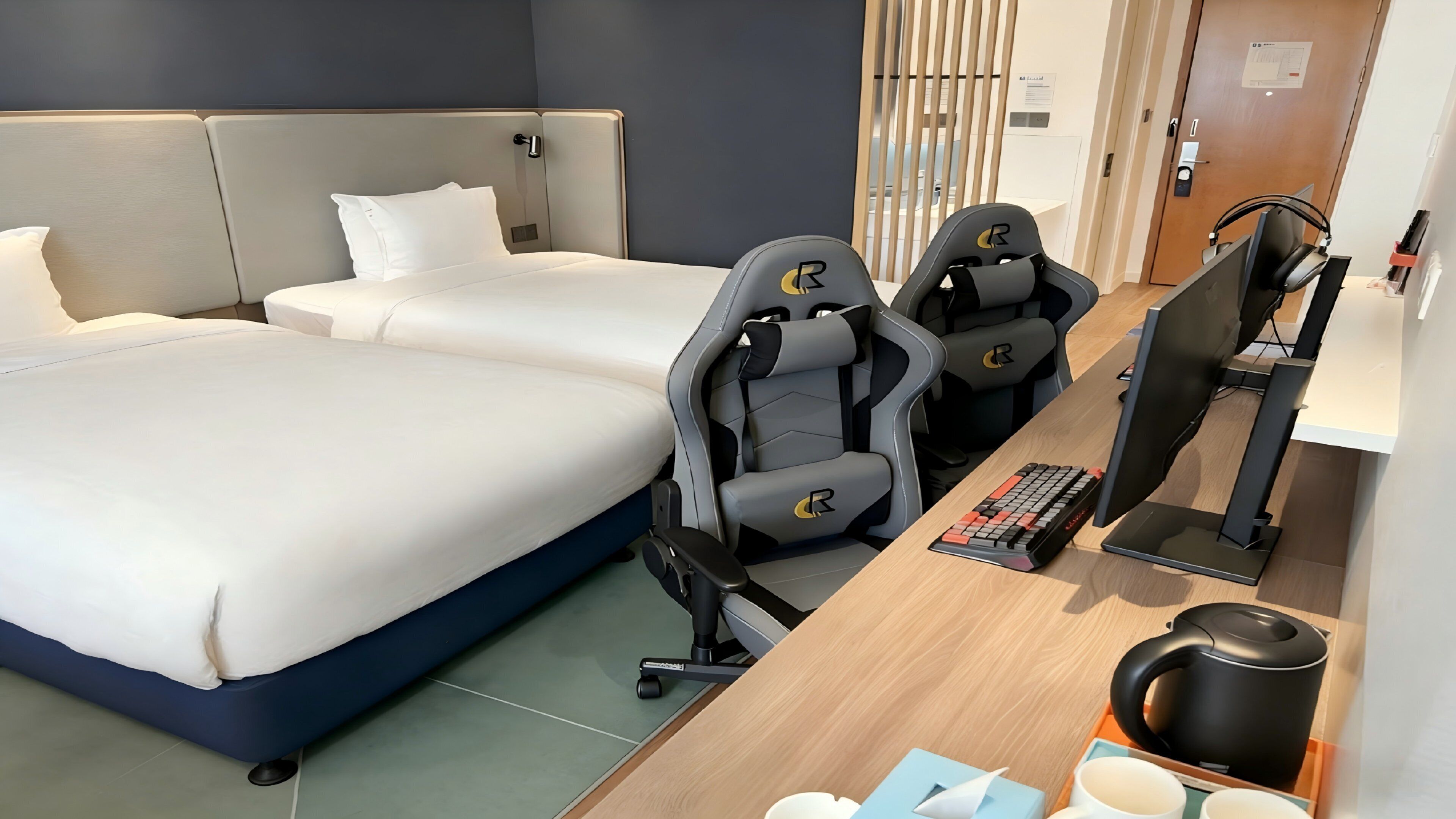 A hotel room with two beds, a desk with a computer, and gaming chairs.